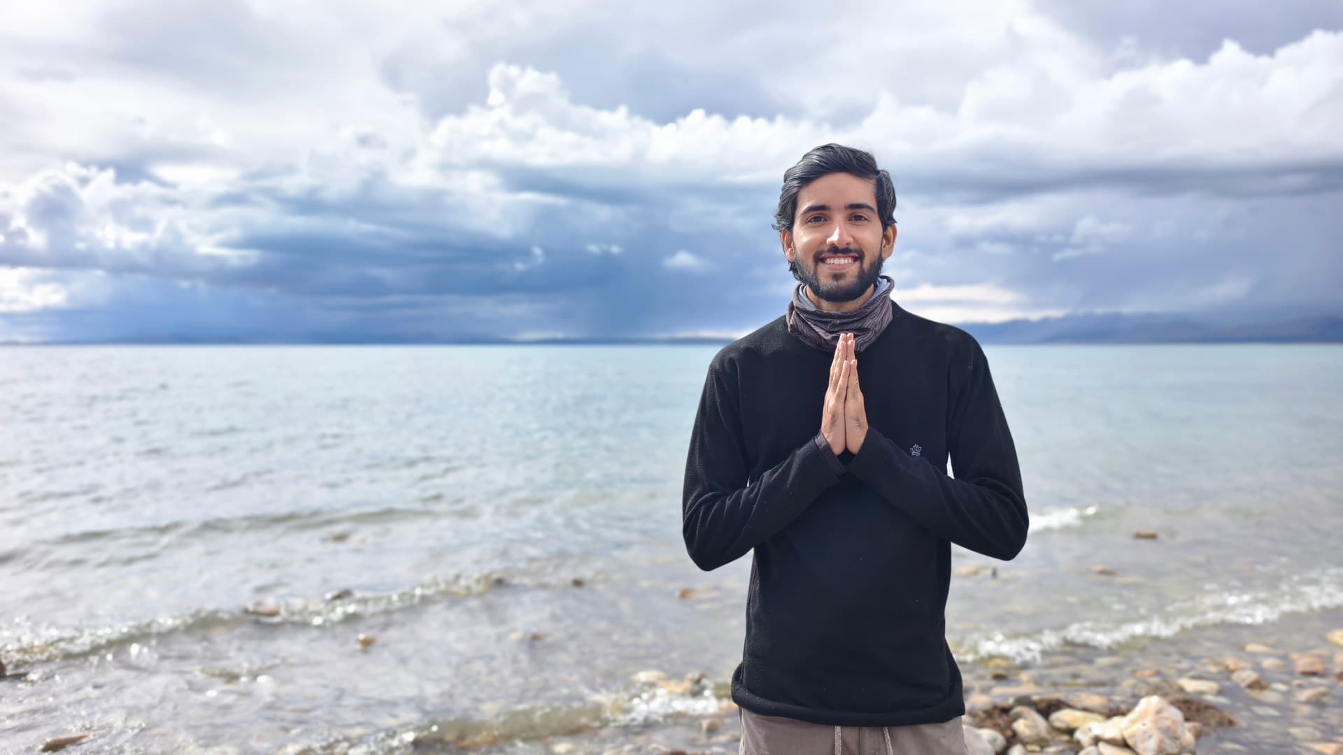 Suryansh at Lake Mansarovar with hands in namaste, mountains and dramatic clouds behind