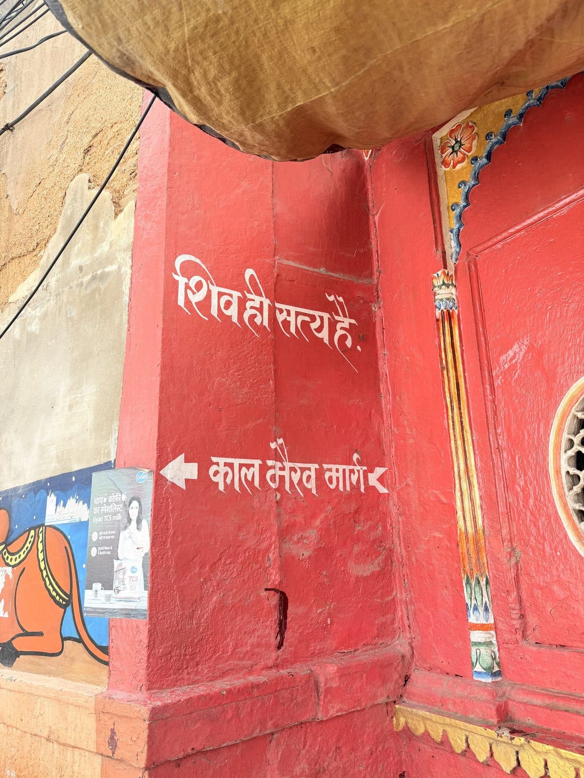 A red wall in a Kashi lane painted with “शिव ही सत्य है” and an arrow marked “काल भैरव मार्ग”, the way to Kaal Bhairav