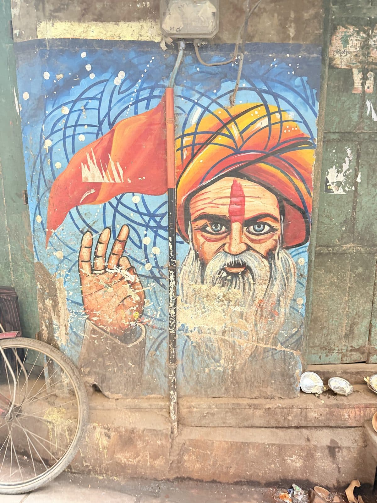 A mural of a bearded sadhu in a saffron turban beside a saffron flag, painted on a weathered lane wall