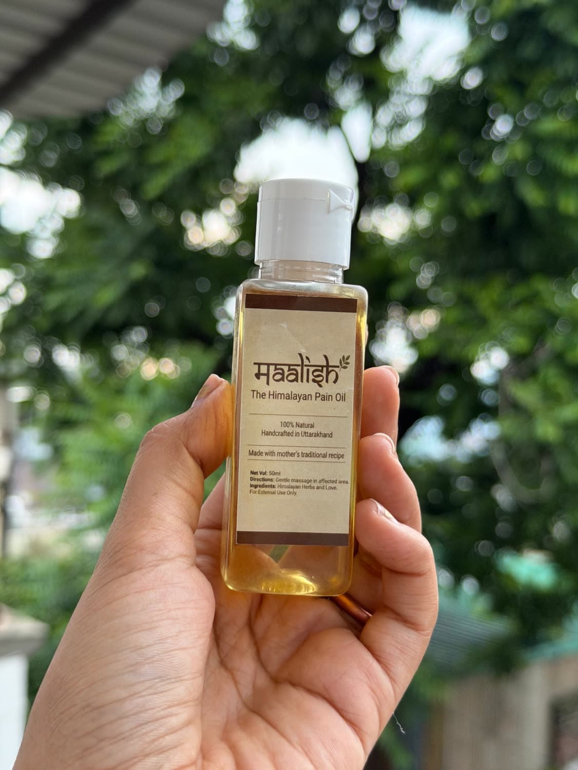A hand holding a bottle of Maalish oil up against sunlit green leaves outdoors.