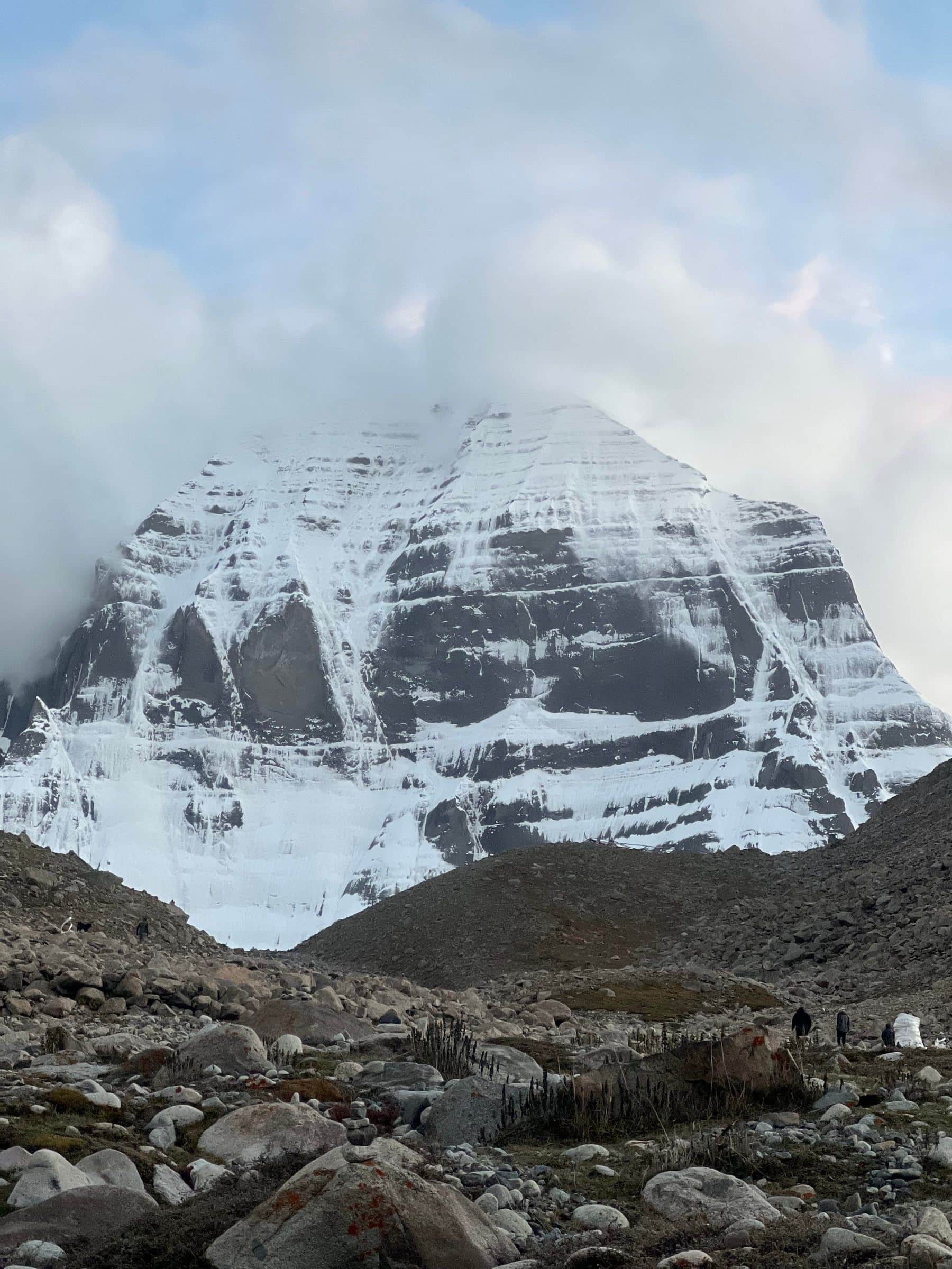 Mount Kailash north face seen from the rocky parikrama trail with pilgrims and a stone cairn in the foreground
