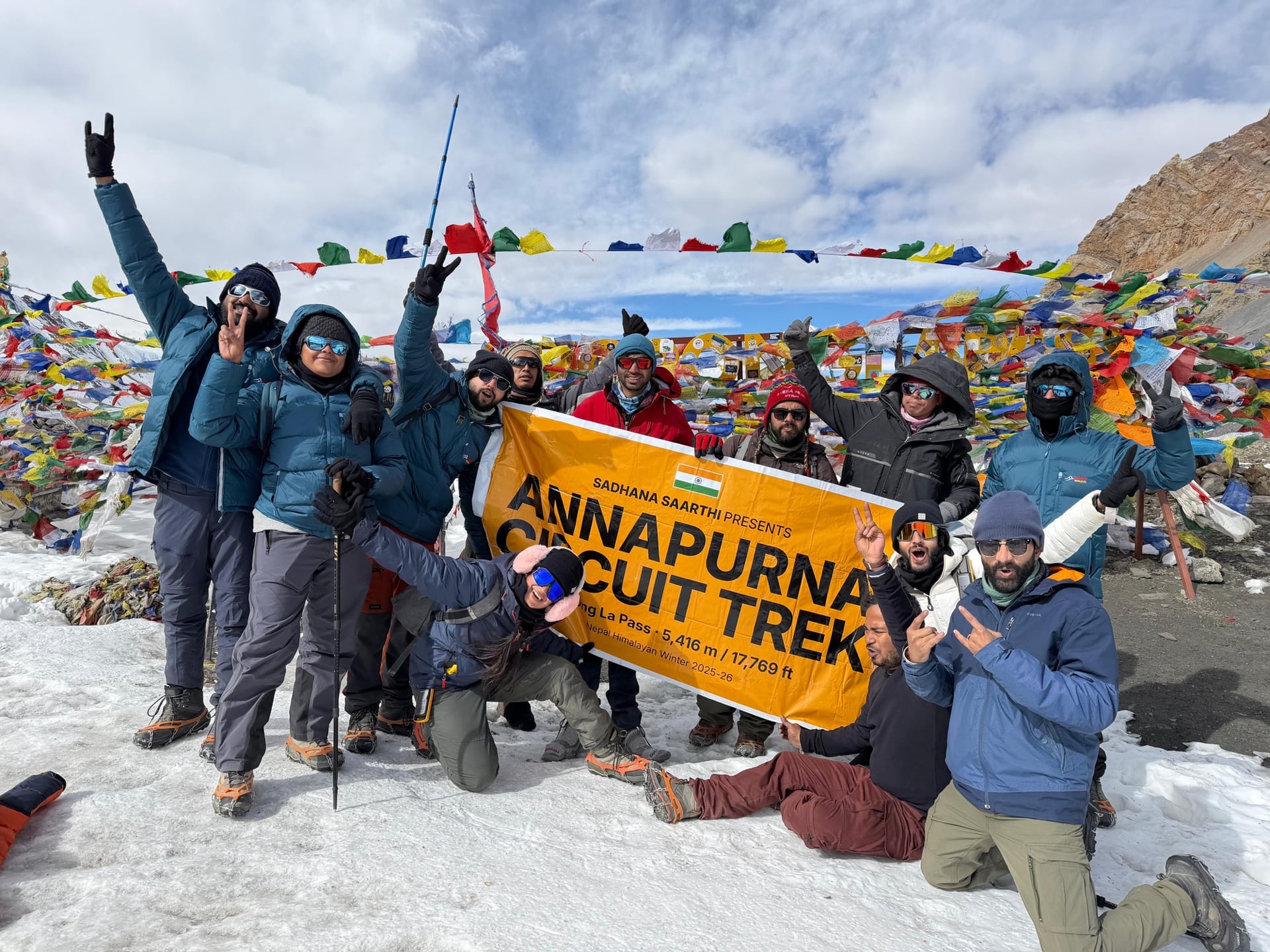 Trekkers at Thorong La Pass on the Annapurna Circuit, with colorful prayer flags and the Himalayas.