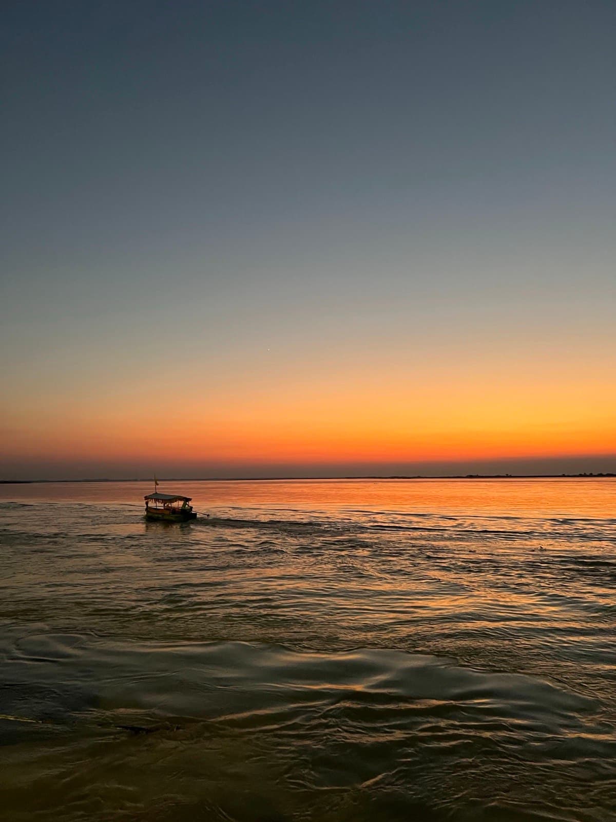 A single boat crossing the wide Ganga at first light, the far bank a thin line under an orange sky